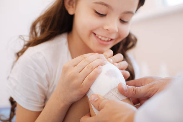 Delighted cute girl looking at her knee Just a scratch. Delighted cute positive girl looking at her knee and smiling while feeling no pain first aid class stock pictures, royalty-free photos & images