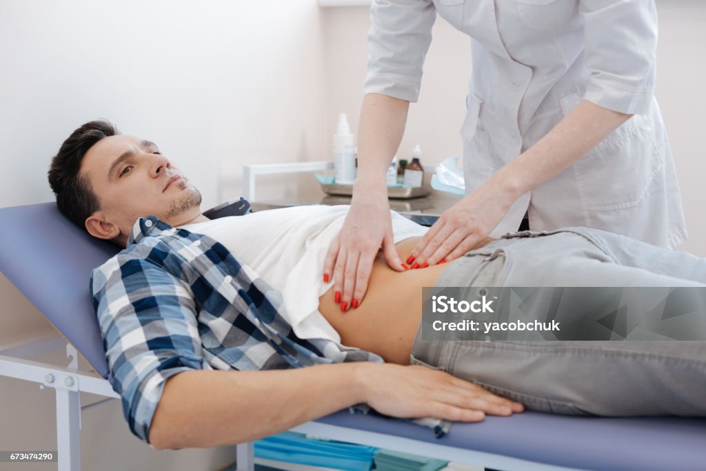 Handsome nice man being examined by the doctor In the clinic. Handsome nice pleasant man lying on the medical bed and being examined by the doctor while being at the hospital Stomachache Stock Photo