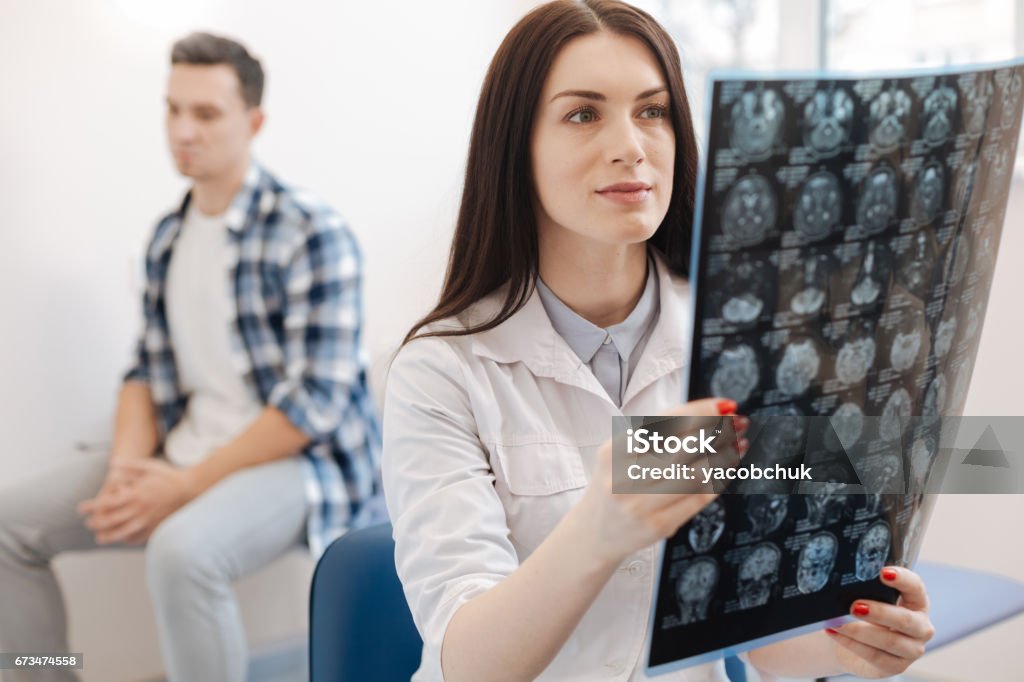 Beautiful attractive doctor holding an X ray photo Visit to the neurologist. Beautiful attractive pleasant doctor holding an X ray photo and looking at while putting a diagnosis Neurologist Stock Photo