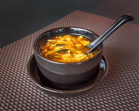 Chinese food, hot and sour soup served in black bowl with spoon on dark background.
