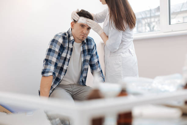 Unhappy sad man receiving the first aid After the accident. Unhappy sad handsome man sitting on the medical bed and receiving the first aid while having the forehead wound concussion photos stock pictures, royalty-free photos & images