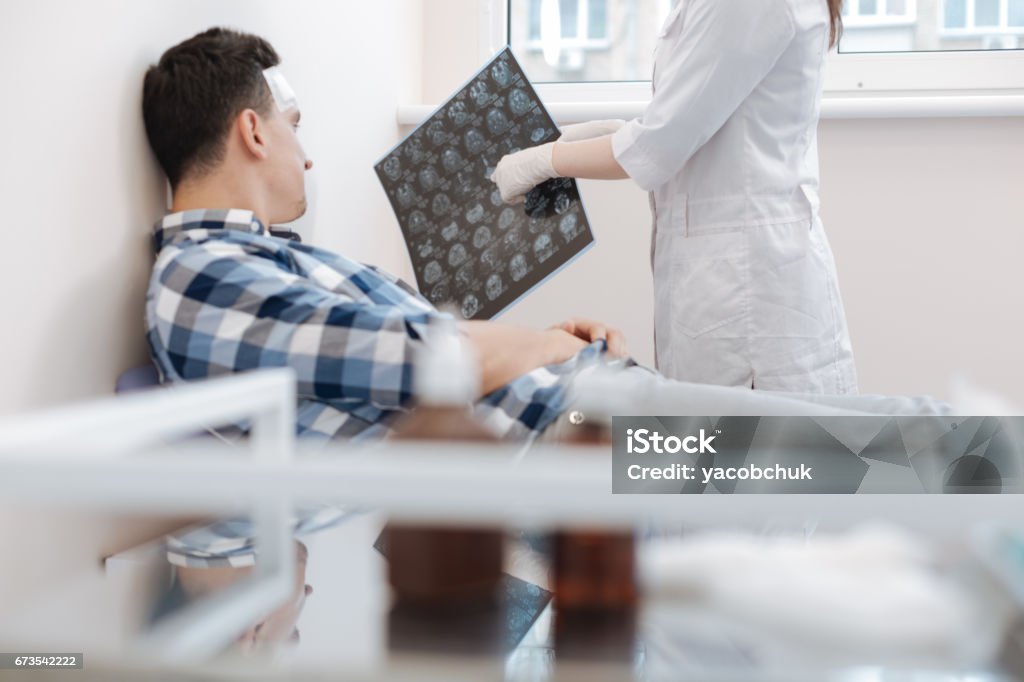 Nice professional doctor pointing at the X ray photo Here is the problem. Nice professional female doctor holding an X ray photo and pointing at it while explaining the patient his problem Recovery Stock Photo