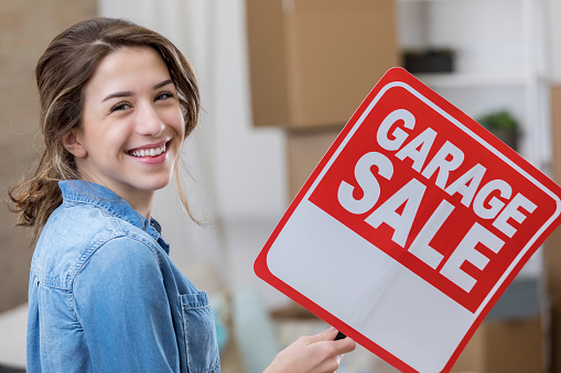 Confident Caucasian young woman holds a garage sale sign. She is preparing to move into a new home or apartment.