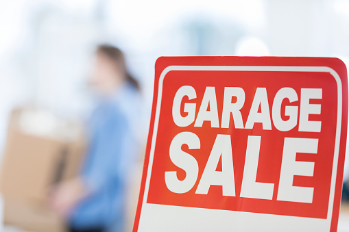 Close up of red garage sale sign. A young woman is packing in the background. Focus is on the sign.