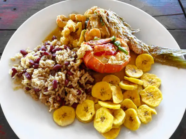 Photo of Local food. Lobster, red snapper fish, shrimp, rice, beans, fried plantains, coconut milk sauce. Roatan Honduras Creole unique traditional cuisine. Delicious seafood lunch, meal rustic wood background