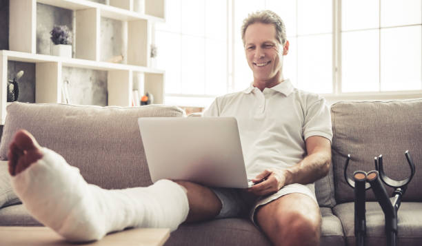 Man with broken leg Handsome mature man with broken leg in gypsum is using a laptop and smiling while sitting on sofa at home man sitting with a bandage on his leg stock pictures, royalty-free photos & images