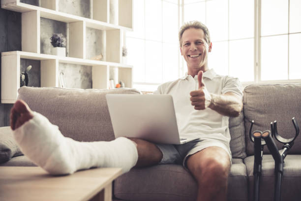 Man with broken leg Handsome mature man with broken leg in gypsum is using a laptop, showing Ok sign and smiling while sitting on sofa at home man sitting with a bandage on his leg stock pictures, royalty-free photos & images