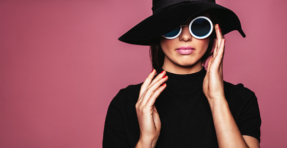 Close up portrait of stylish caucasian female model. Beautiful woman in hat and sunglasses posing over pink background.