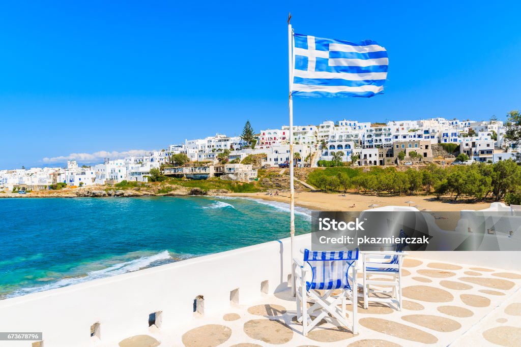 Greek flag waving on a terrace overlooking Piperi beach in Naoussa village, Paros island, Cyclades, Greece The island of Paros is one of the most famous Greek islands of the Aegean Sea and it belongs to the Cyclades islands archipelago. Apartment Stock Photo