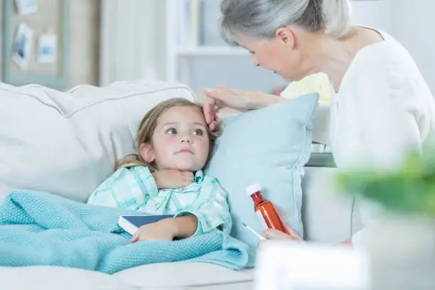 Caring Caucasian grandmother cares for her ill granddaughter. The woman is holding a bottle of prescription medication while checking the girl for a fever.