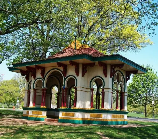 Colorful structure, created with Asian architecture/design, used as a shelter. This pavillion is located in Druid Hill Park (Baltimore, MD) at the top of a hill, alongside a street that runs throughout the park, situated beneath a group of trees.
