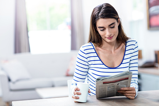 Young woman smiling while reading newspaper by table at home