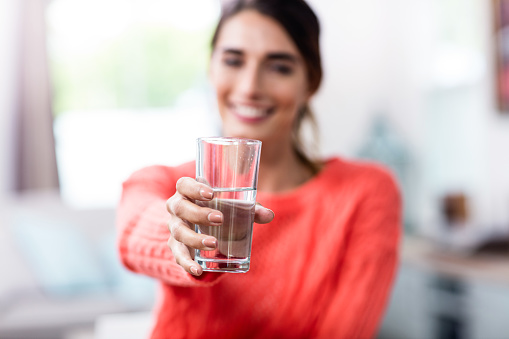 Happy young woman showing drinking glass with water at home