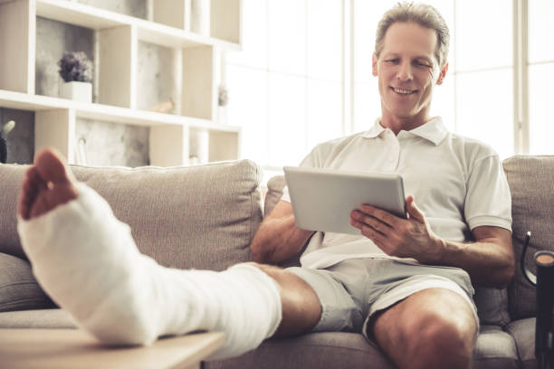 Man with broken leg Handsome mature man with broken leg in gypsum is using a digital tablet and smiling while sitting on sofa at home man sitting with a bandage on his leg stock pictures, royalty-free photos & images