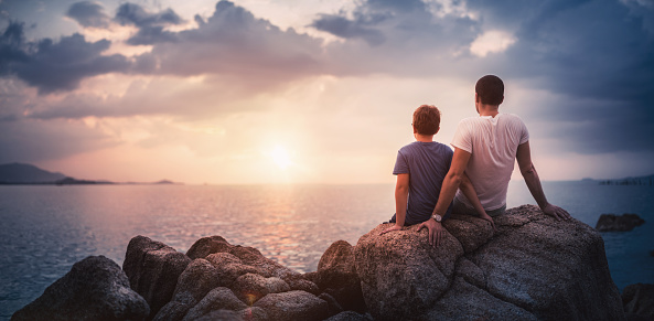 Father with son sitting on rocks and watching the sunset over sea. Stock photo.