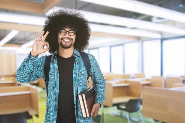 College student showing ok sign while holding book and standing in the classroom