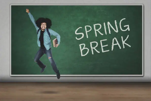 College student holding book while leaping with spring break text on the chalkboard