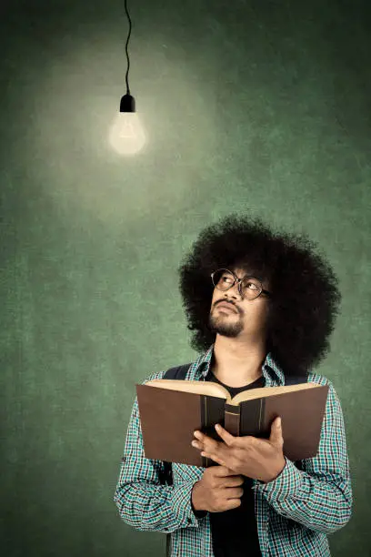Young college student thinking idea while holding a book with light bulb over his head