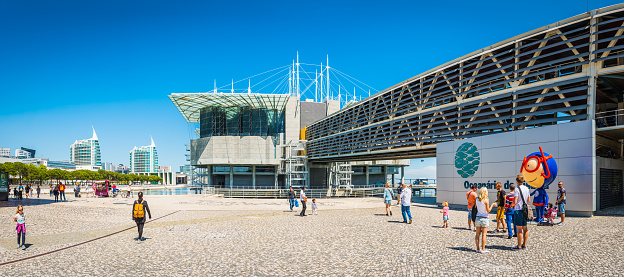 Families of tourists and locals enjoying the sunshine outside the Oceanario de Lisboa in the modern waterfront development of Parque das Nacoes in Lisbon, Portugal.