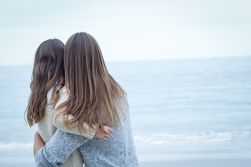 Rear view of mother embracing daughter at sea shore against sky