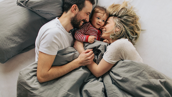 Photo of young parents enjoying morning routine in a cosy bed with their baby boy