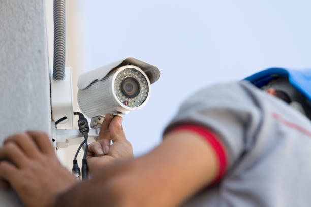A technician in a gray shirt with a red trim adjusts a security camera mounted on a wall, focusing on the camera's lens while wearing a blue hard hat.