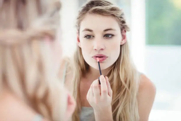 Young woman applying lip gloss while looking in mirror