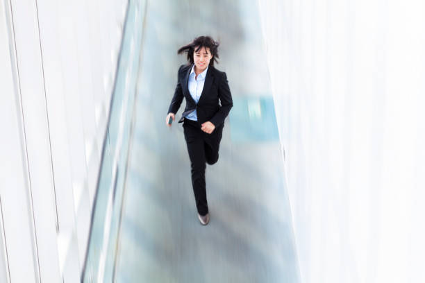 A young Japanese businesswoman is in a rush and running on a flat floor while holding a smartphone.