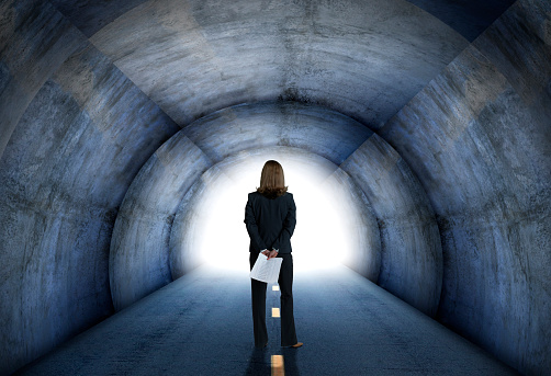 A businesswoman holds some paperwork behind her back as she looks towards a bright light at the end of a tunnel that she is standing in.