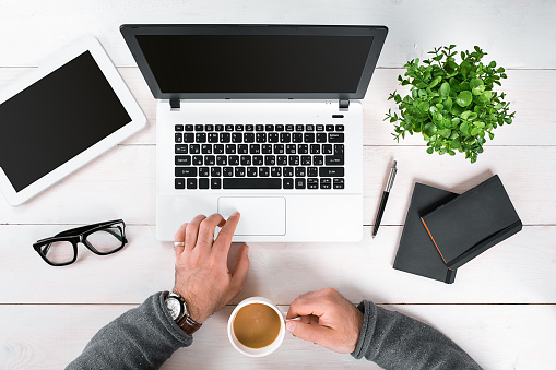 Directly above view of human hands typing on laptop. Laptop, digital tablet, diary, coffee cup and potted plant on work desk. Man working from home.