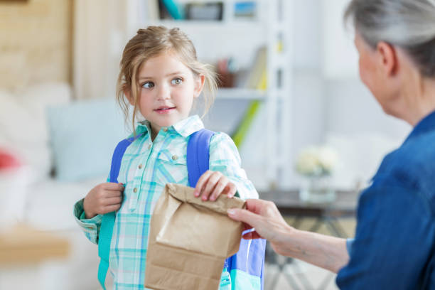 Grandma gives granddaughter a sack lunch Grandmother hands elementary age granddaughter a packed lunch as the girl leaves for school. The girl is wearing a backpack. bag lunch stock pictures, royalty-free photos & images