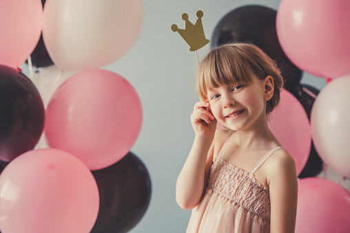 Cute little princess in dress is holding a crown, looking at camera and smiling, on gray background with colorful balloons