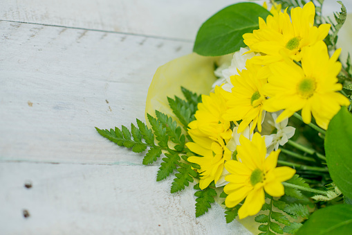 A bouquet of yellow and white daisies with leather fern wrapped in yellow paper rests on a white wooden background. It is a Mother's day gift.