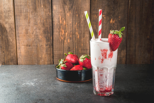 Milkshake with strawberries. In a glass on a stone black and dark wooden background. With strawberries in a bowl next to and striped bright straws. copy space