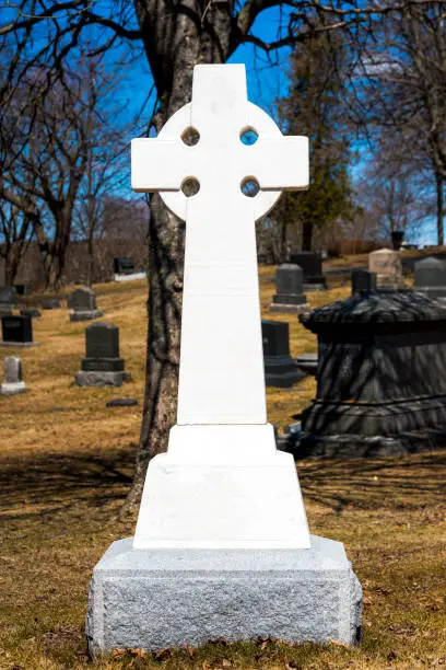 Bright white marble Celtic cross in a dark graveyard. Trees and grave markers behind. Blue sky behind the trees. No text on the cross.