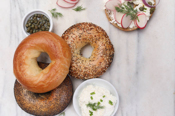 An overhead close up horizontal photograph of three assorted bagels, two small containers of capers and cream cheese, sliced radishes and half a bagel all ready to go.