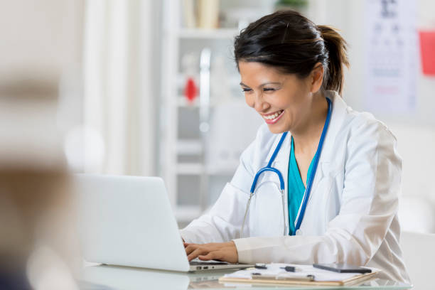 улыбающийся педиатр использует компьютер в своем кабинете - laptop doctor using computer nurse стоковые фото и изображения