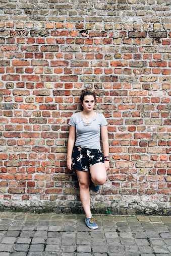 Full length portrait of serious expression young adult woman against brick wall.