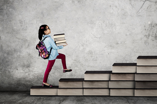 Picture of a little school girl carrying a pile of books while walking on the stair made of books. Back to School concept