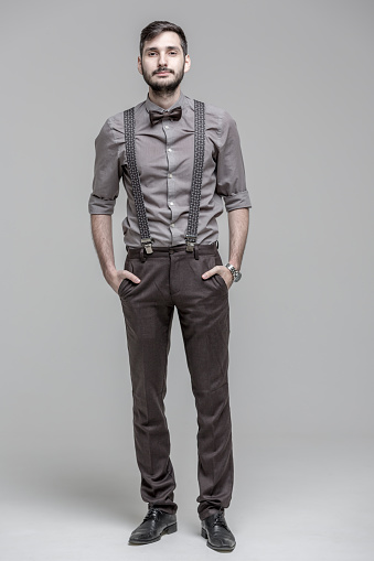 Full-length portrait of handsome bearded man dressed in men's classic clothes (shirt, trousers with suspenders and bow tie). The man is standing with hands in pants pockets. He is calmly looking at the camera. Studio shooting on light gray background