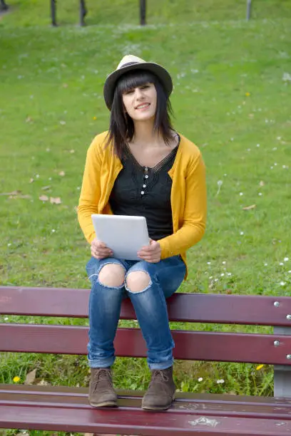 portrait of young brunette girl sitting on a bench with a tablet in park, outside