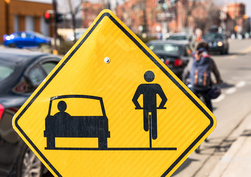 Close-up of a sign advising  drivers and cyclists to share the road, with cars and a cyclist defocused in the distance.