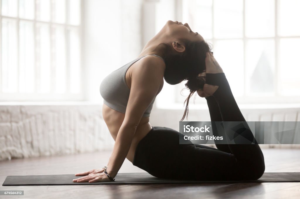 Young attractive woman in King Cobra pose, loft studio backgroun Young yogi woman practicing yoga concept, doing King Cobra exercise, Raja bhudjangasana pose, working out wearing black sportswear, full length silhouette on white loft studio background. Side view Adult Stock Photo