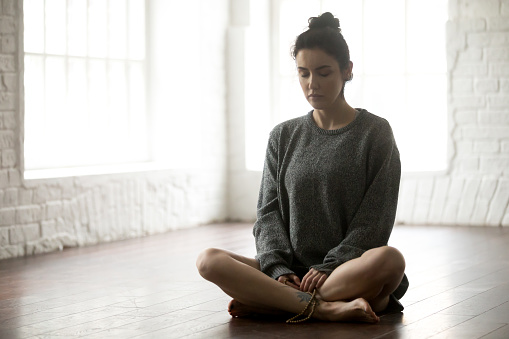 Young calm yogi woman practicing yoga concept, sitting in Sukhasana exercise, Easy Seat pose, her eyes closed, working out wearing grey oversized sweater, beads in hands, full length studio background