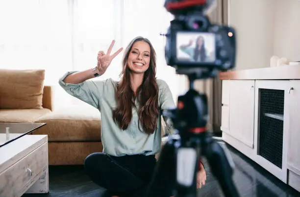 Young woman recording her daily video blog on a tripod mounted camera. Smiling young woman showing victory or peace sign on camera.