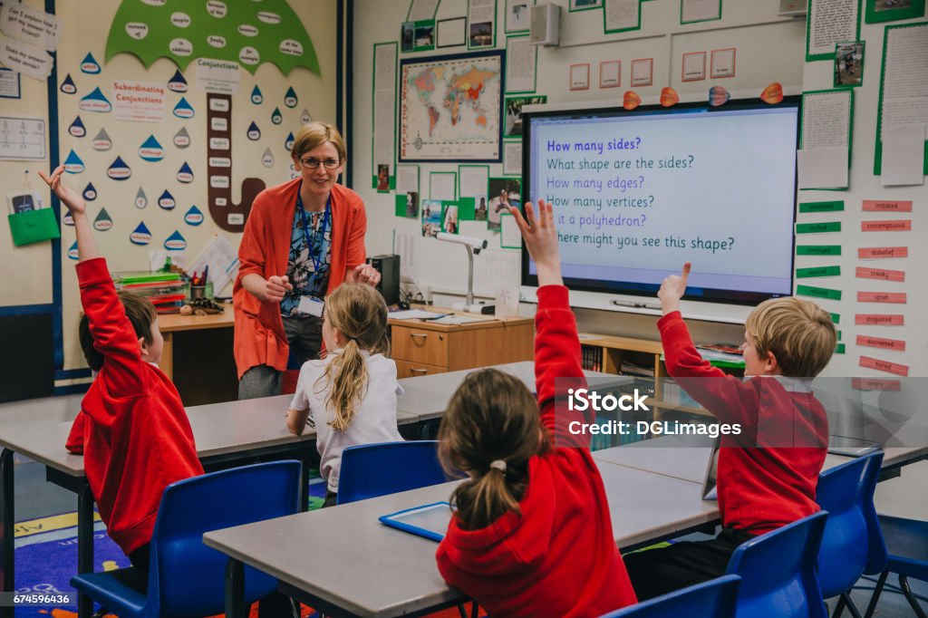 Primary School Lesson Female teacher is teaching shapes to her primary school students. She is asking hem a question and some of the students have their hand in the air to answer. Teacher Stock Photo