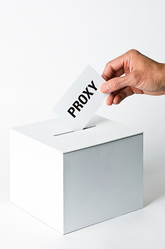 A hand dropping a proxy vote into a proxy ballot box. Photographed on white background.