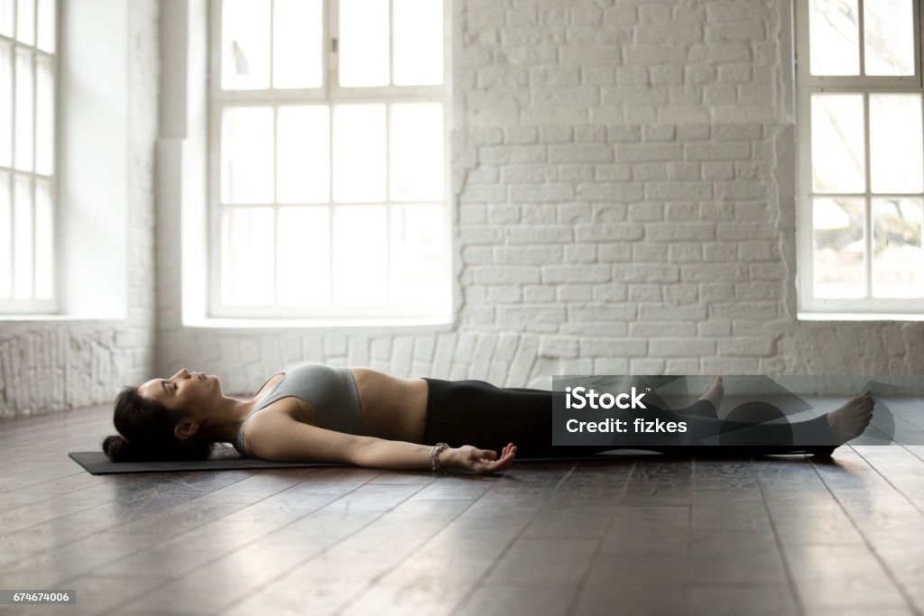 Young attractive woman in Savasana pose, white loft studio backg Young attractive yogi woman practicing yoga exercises concept, lying in Dead Body, Savasana, Corpse pose, resting after working out, wearing sportswear, full length, white loft studio background Lying On Back Stock Photo