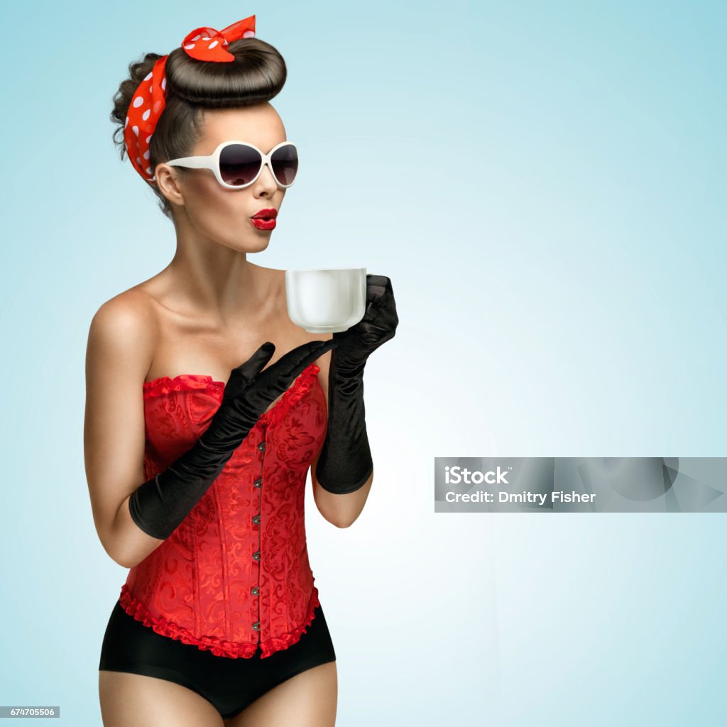 Hot beverage. Three-quarter portrait of glamourous pinup girl wearing vintage gloves and red ribbon in her hair, holding a cup of hot coffee or tea and cooling it. Adult Stock Photo