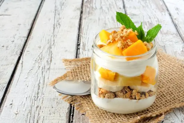 Tropical mango and pineapple parfait in a mason jar on a rustic white wood background